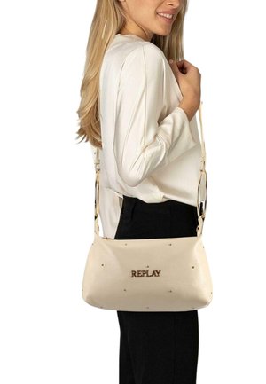 Mujer con blusa blanca y pantalones negros que lleva un bolso de hombro beige de Replay con pequeños adornos y herrajes dorados.