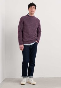 Bordo adīts džemperis ar augstu kaklu, valkāts virs gaiši zilas krekla. Apvienots ar tumšiem džinsu biksēm un krēmkrāsas kedām.