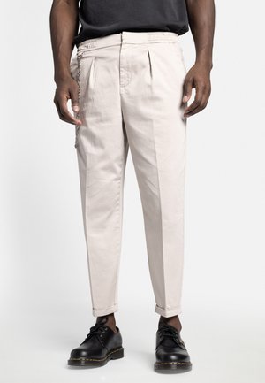 Man med beige plisserade byxor med uppvikta fållar, svarta läderskor och en kedjeaccessoar på vänster sida framför en enkl vit bakgrund.