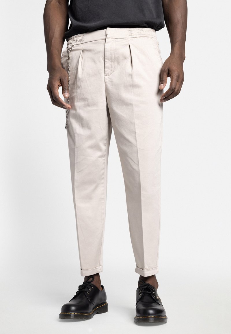 Man med beige plisserade byxor med uppvikta fållar, svarta läderskor och en kedjeaccessoar på vänster sida framför en enkl vit bakgrund.