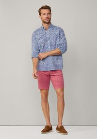 Camisa azul con estampado y cuello mao, llevada con pantalones cortos rosas y mocasines de ante marrón. La camisa tiene mangas largas y un diseño geométrico.