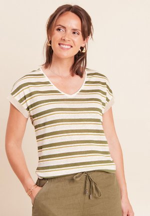 Frau lächelt, trägt ein weiß-olivgrün gestreiftes kurzärmliges Shirt mit olivgrünen Hosen mit Kordelzug, accessoriert mit goldenen Ohrringen und Armbändern.