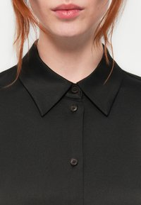 Svart skjorte med krage i en glatt tekstur, med knappestopp og rund krage. Nærbilde som fremhever stoffet og knappene.