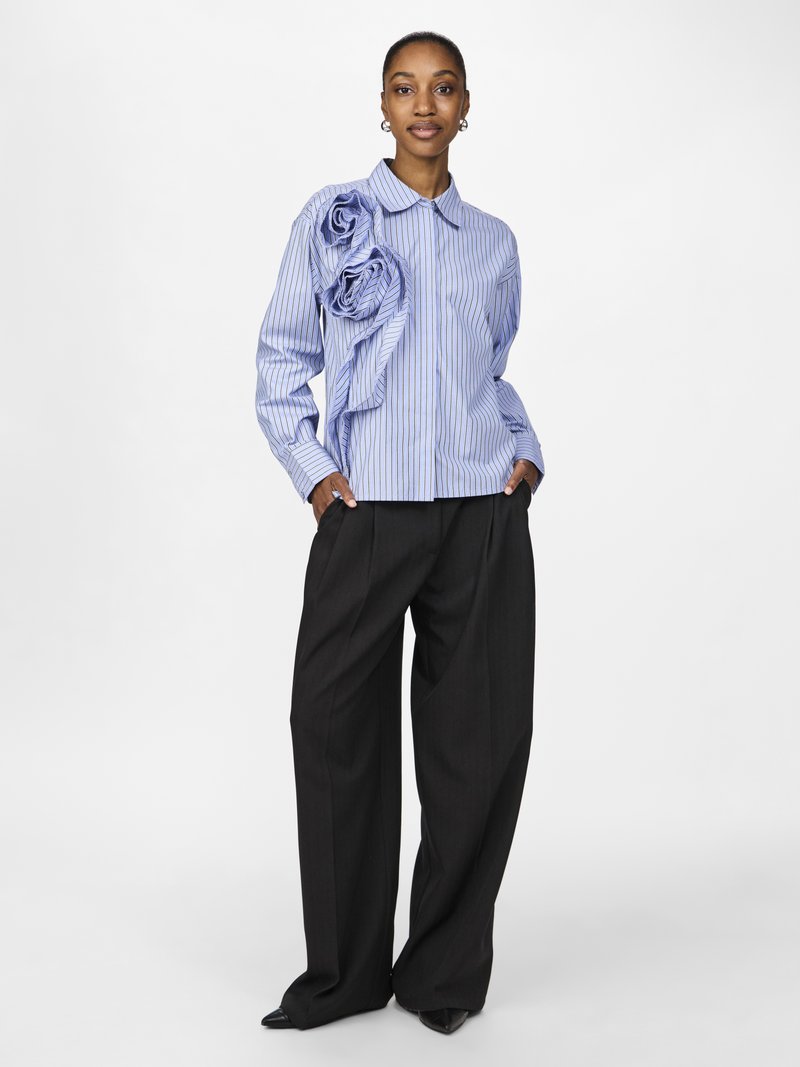 Blauw-wit gestreepte shirt met grote, zacht aanvoelende rozen van textiel op de linkerschouder, gecombineerd met zwarte wijde broek en zwarte puntige schoenen.