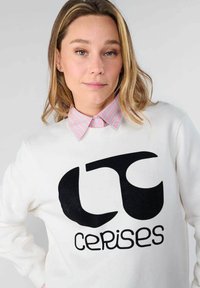 Sudadera blanca con un gran logo gráfico negro y texto "cerises". Se puede ver un colorido cuello a rayas rosa y blanco por debajo.