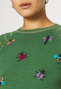 Suéter verde de punto con insectos bordados en naranja, azul, rojo, negro y morado. Presenta un cuello redondo y costuras con acento dorado.