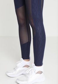 Marina leggings med en texturerad diamantskiss och genomskinliga paneler, som bärs med vita sportskor med nätdetaljer.