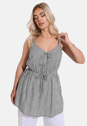 Lyshåret kvinde iført ærmeløs sort-hvid gingham-kjole med bindebånd ved udskæring og talje, kombineret med hvide bukser, poserer foran hvid baggrund.