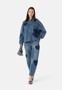 Denimjakke og bukse i medium blå med frynsete kanter, med store marineblå paljett-hjertepatcher. Modellen holder en vevd grå håndveske.