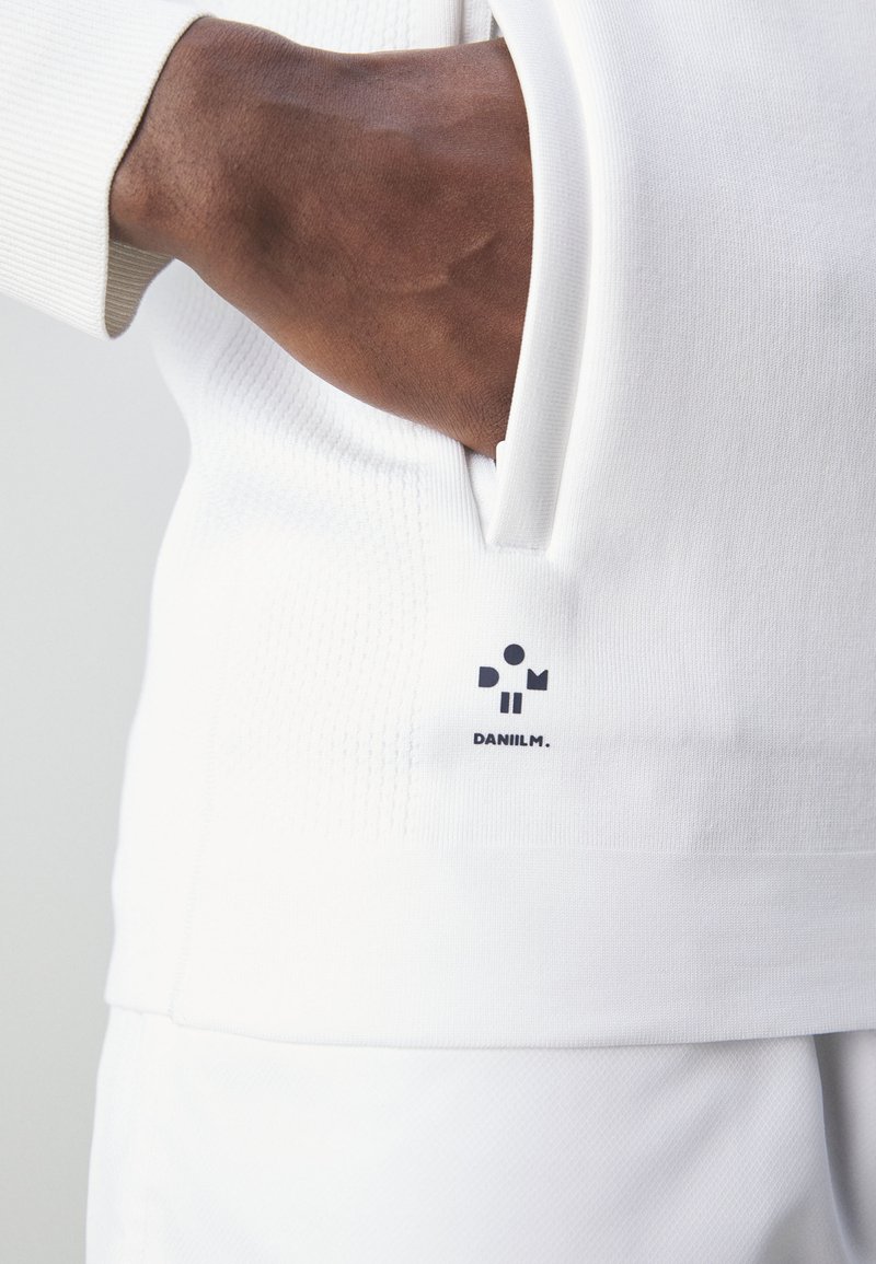 Sweatshirt blanc avec une texture côtelée, comportant une poche latérale et une étiquette logo noire portant "DANILM." en bas à gauche.