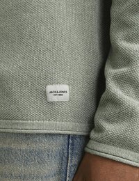 Hellgrüner, gestrickter Pullover mit strukturierter Oberfläche, mit einem Markenetikett am Saum und gerippten Bündchen. Kombiniert mit ausgewaschenen Jeans.