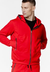 Czerwona bluza z kapturem na zamek błyskawiczny, z czarnym wnętrzem, przednimi kieszeniami, ściągaczami przy mankietach i kapturem ze sznurkiem. Gładka tekstura i sportowy krój.