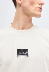 Νέος άντρας που φοράει λευκό πουκάμισο με μια μαύρη ορθογώνια ετικέτα που γράφει "HUGO" στο στήθος, με το κεφάλι γυρισμένο προς τα δεξιά.