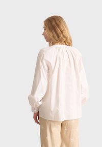 Weißes Langarmhemd mit gerafftem Rückendetail, aus leichtem Stoff, kombiniert mit beigen, gemusterten Hosen.