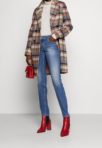 Plaid coat in brown, beige, and navy, paired with a light sweater, blue jeans, and red heeled ankle boots. Small red handbag included.