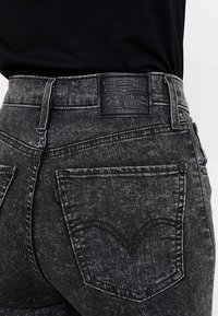 Los vaqueros de mezclilla negra cuentan con una cintura alta, dos bolsillos traseros con costuras distintivas y una etiqueta de cuero de la marca en la pretina.