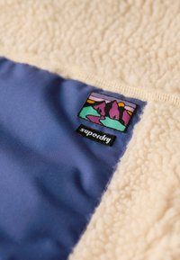 Tissu en polaire crème associé à un panneau en satin bleu foncé. Présente un patch de marque coloré représentant des montagnes et le texte "superdry".