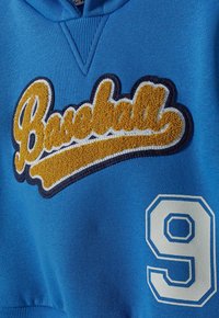 Blå huvtröja med upphöjd gul broderad text "Baseball" och ett vitt, konturerat nummer 9 på den nedre sidan.