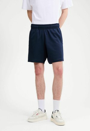 Uomo che indossa pantaloncini elasticizzati blu navy, calzini bianchi alti, sneakers bianche con dettagli verdi e rossi, e una maglietta bianca, in piedi.