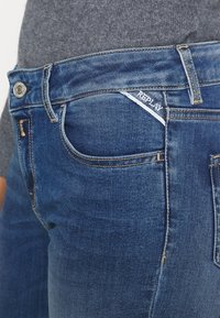 Jean en denim bleu foncé avec une poche avant arborant un petit label « Replay ». Le tissu a une texture lisse et des détails de couture visibles.