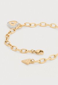 Βραχιόλι από μέταλλο σε χρυσό τόνο με οβάλ συνδέσμους, κλείδωμα σε σχήμα καρδιάς με στρας και τρίγωνο μενταγιόν με χαρακτική τη λέξη «GUESS».