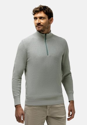 Man draagt een lichtgrijze trui met rits en beige broek, staat met ontspannen handen en kijkt naar links tegen een effen achtergrond.