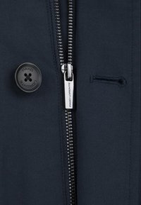 Granatowa kurtka z bliska, z metalowym zamkiem błyskawicznym, okrągłym czarnym guzikiem z logo "PAKO LORENTE" oraz małym, zaszewkowanym detalem kieszeni.