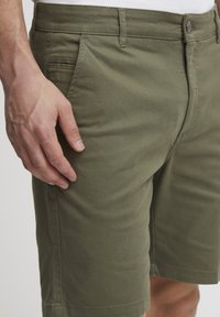 Olivenfargede shorts i en bomullsblanding med en jevn tekstur, med sidelommer og knappelukking. Forsiktig sydd sømmer er synlige.