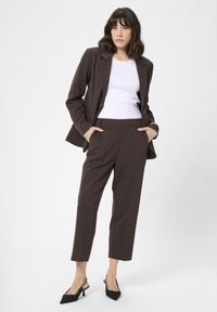 Brauner maßgeschneiderter Blazer mit passender Hose, darunter ein weißes, figurbetontes Oberteil und schwarze Slingback-Absätze; glatter Stoff mit strukturiertem Design.