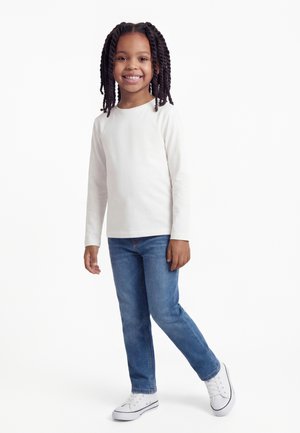 Bambino sorridente con i capelli intrecciati, in piedi, indossa una maglia bianca a maniche lunghe, jeans blu e scarpe da ginnastica bianche su uno sfondo bianco semplice.