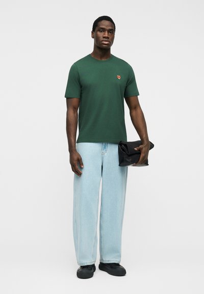 Άνδρας που φοράει πράσινο κοντομάνικο μπλουζάκι με μικρό έμβλημα αλεπούς, ανοιχτού μπλε χρώματος παντελόνι με φαρδύ πόδι, μαύρα παπούτσια, κρατώντας μαύρο διπλωμένο τσαντάκι χειρός.