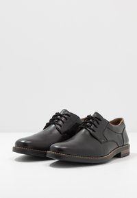 Scarpe di pelle nera con una superficie liscia, punta rotonda e accenti marroni a contrasto sulla suola e sul tallone. Presentano lacci tradizionali.