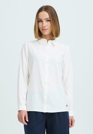 Mujer con cabello hasta los hombros, vestida con una camisa blanca abotonada y pantalones oscuros, de pie frente a un fondo claro y sencillo.