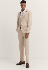 Beige maßgeschneiderter Anzug mit einem taillierten Blazer und schmalen Hosen, kombiniert mit einem weißen Hemd und braunen Slippern. Subtile Texturen und klare Linien.