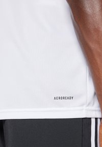 Camiseta deportiva Aeroready blanca con tejido texturizado, de mangas cortas y con un logotipo impreso cerca del dobladillo. Se lleva con pantalones cortos oscuros con rayas.