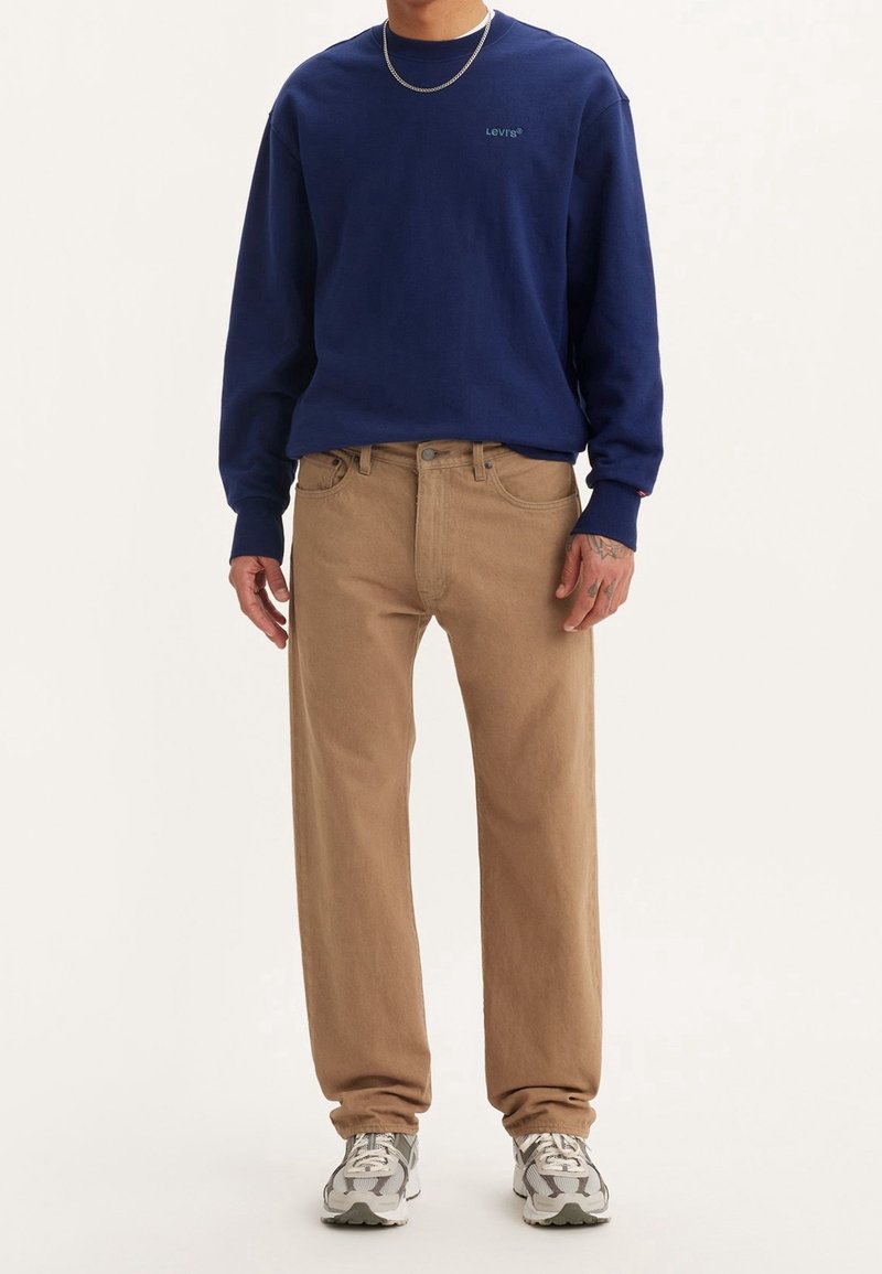 Homme portant un sweat-shirt Levi's bleu marine, un pantalon beige droit, des baskets blanches et grises, et un collier chaîne argenté.