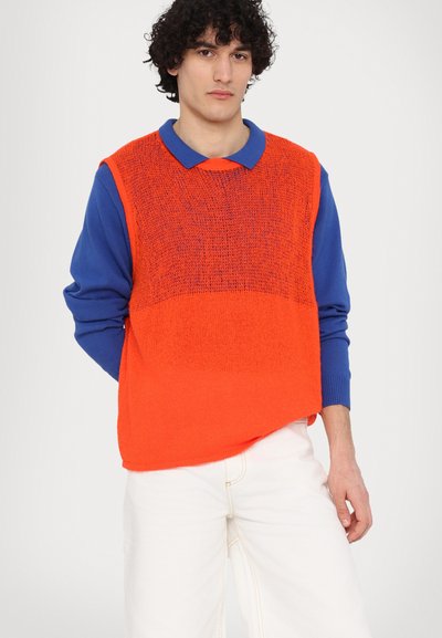 Chaleco de punto naranja con un diseño holgado, superpuesto a una camisa de manga larga azul, acompañado de pantalones blancos. Textura suave y colores contrastantes.