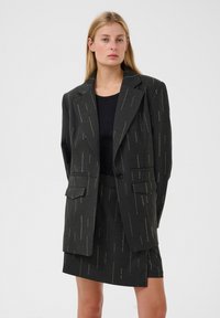 Sötétszürke zakó világos függőleges csíkokkal, két elülső zsebbel és egy gombos zárással, párosítva egy azonos anyagú szoknyával.