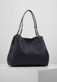Sac à main en cuir noir avec texture granuleuse, chaîne argentée et doubles poignées en cuir, posé sur un socle blanc devant un fond gris clair.
