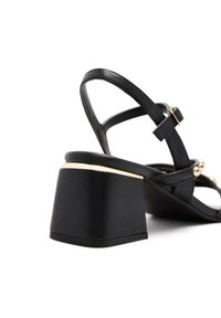 Sandalias negras de tacón alto con tacón cuadrado, que presentan múltiples correas delgadas y acentos en color dorado. Superficie textil suave con un detalle mínimo.