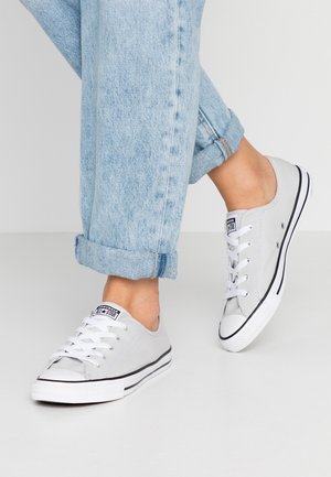 Henkilö, jolla on käännetyt vaaleansiniset farkut ja valkoiset matalat Converse All Star -tossut, seisoo yksinkertaisella vaaleanharmaalla lattialla.