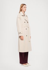 Beiger Trenchcoat mit doppelreihigem Design, großem Kragen und geknöpften Manschetten. Ein taillierter Gürtel und seitliche Taschen erhöhen die Funktionalität.
