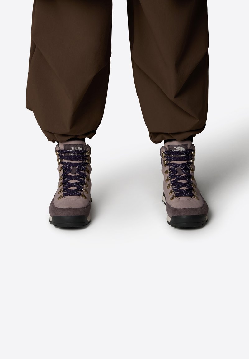 Botas de senderismo en púrpura y gris que cuentan con parte superior de ante y textil, ojales dorados y cordones azules, con una suela gruesa de goma negra.