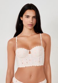 Bra in pizzo rosa chiaro con bordi scallop, ricami floreali, spalline regolabili e un dettaglio centrale con ritaglio. Design strutturato, stile aderente.