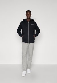 Schwarze Zip-Hoodie mit Kordelzügen und Fronttaschen, kombiniert mit hellgrauen Jogginghosen mit Logo-Akzenten, abgerundet mit weißen Sneakern.
