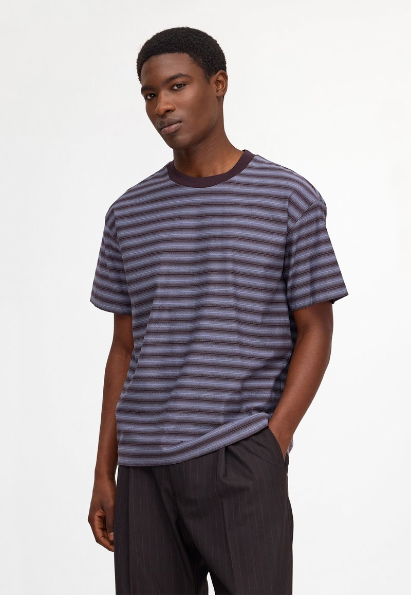Jeune homme portant un t-shirt rayé horizontal bleu et noir et un pantalon sombre à fines rayures, une main dans la poche.
