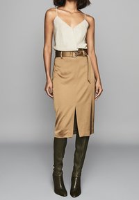 Vrouw in een mouwloze beige top, hoge camel rokje met een split aan de voorkant en een riem, gecombineerd met donkergroene kniehoge laarzen.