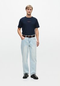 Ναυτικό βαμβακερό t-shirt με κόκκινη εκτύπωση, ανοιχτό μπλε τζιν χαλαρής γραμμής, καφέ δερμάτινη ζώνη και μαύρα παπούτσια, στέκεται σε απλό φόντο.