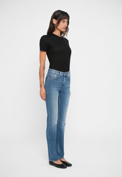 Jeans de mezclilla azul con corte recto y un ligero desvanecido, combinados con una blusa negra ajustada de manga corta y zapatos planos negros.