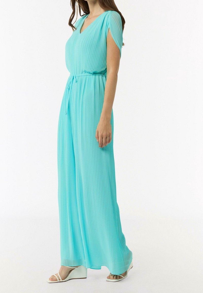 Femme portant une combinaison turquoise plissée sans manches avec jambes larges et sandales compensées blanches, debout devant un fond blanc.